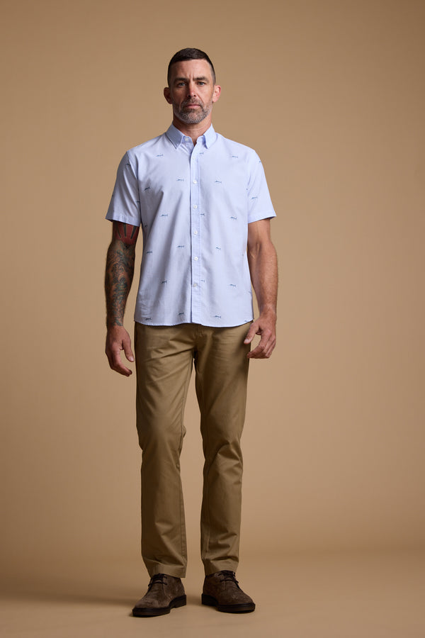 A man with short dark hair, a trimmed beard, and tattoos on his left arm stands against a beige background wearing the Barkers Marlin Stripe Shirt and khaki pants with brown shoes.