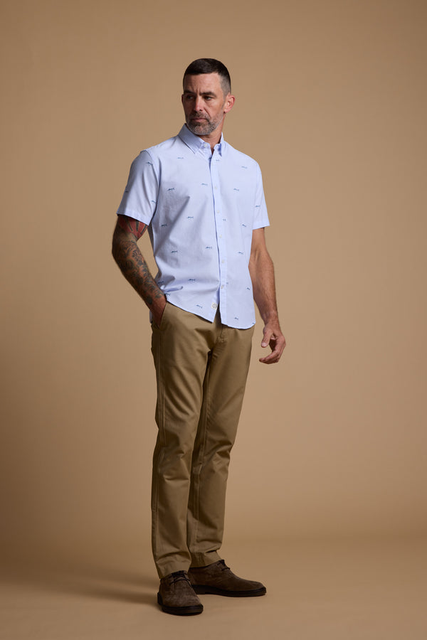 A man with short hair and tattoos on his right arm stands against a tan background, wearing the Barkers Marlin Stripe Shirt made from responsibly sourced cotton, khaki pants, and brown shoes.