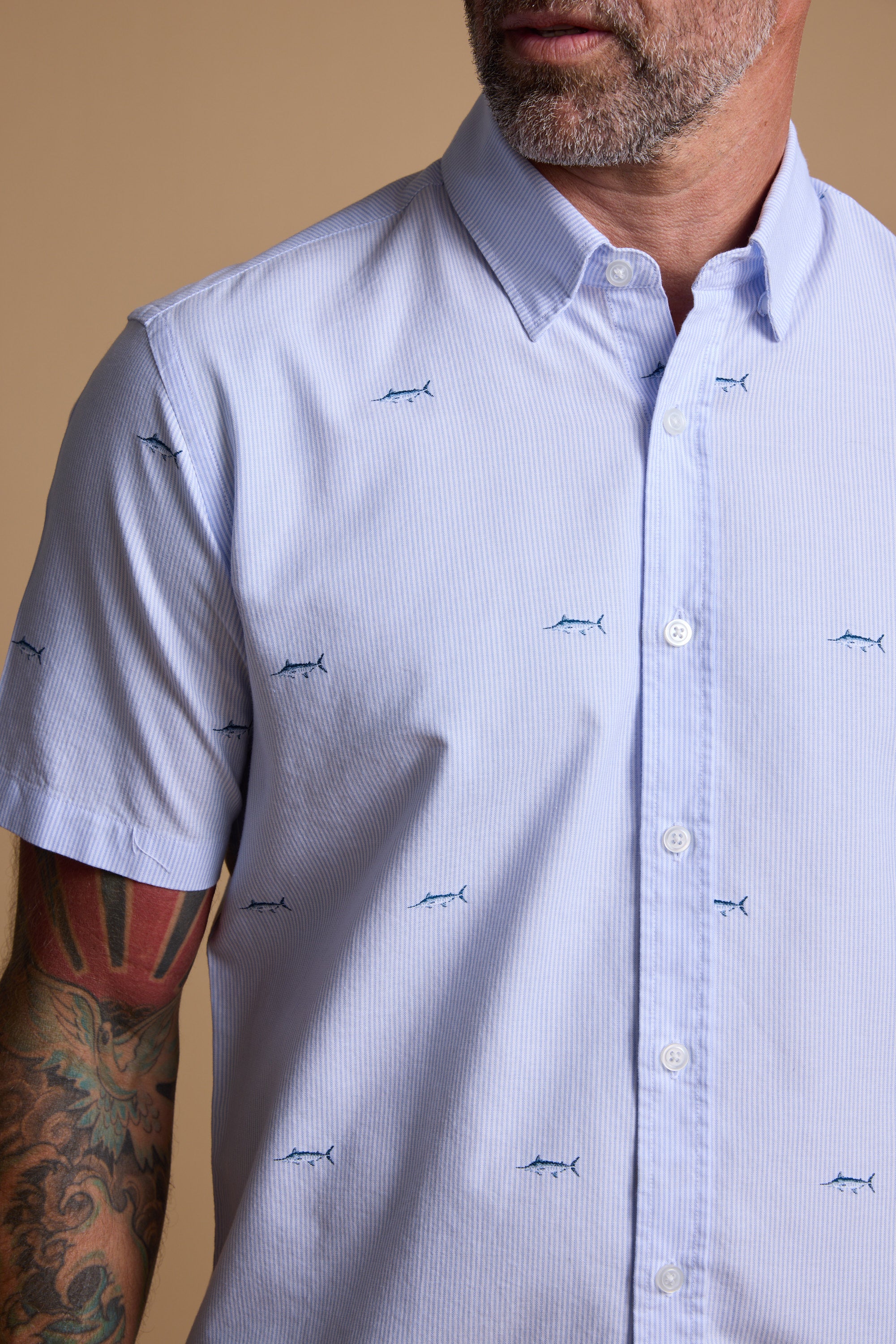A man with a gray beard and a colorful tattoo on his left arm wears the Barkers Marlin Stripe Shirt, crafted from responsibly sourced cotton. The shirt is light blue with embroidery. He stands against a plain beige background.