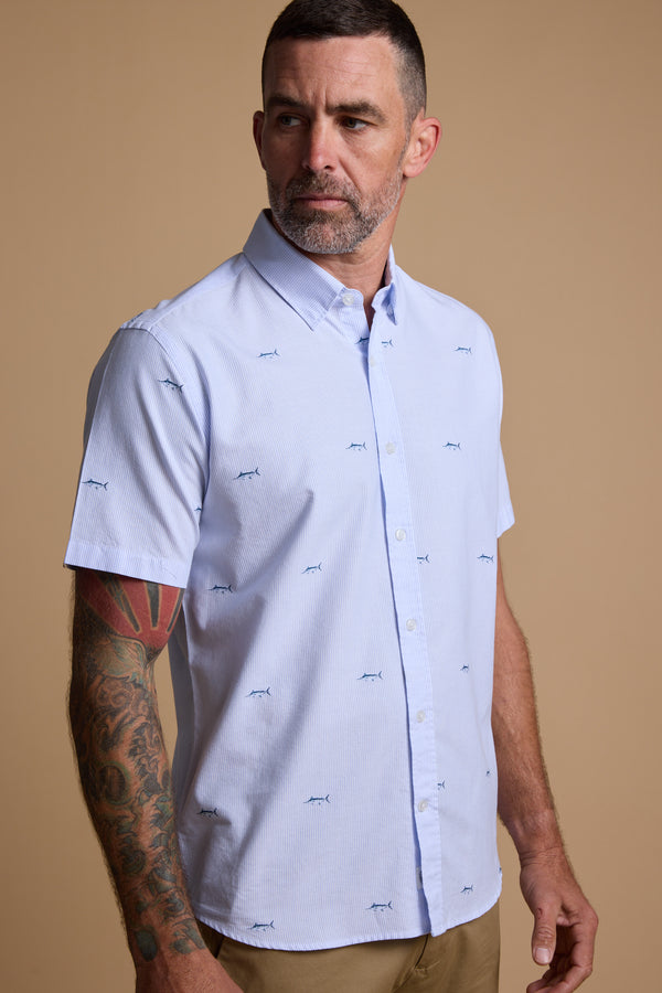 A man with short hair and a beard wears the Barkers Marlin Stripe Shirt, a light blue, responsibly sourced cotton button-up with blue shark prints, and tan pants. He has a sleeve tattoo on his left arm and stands against a plain beige background.