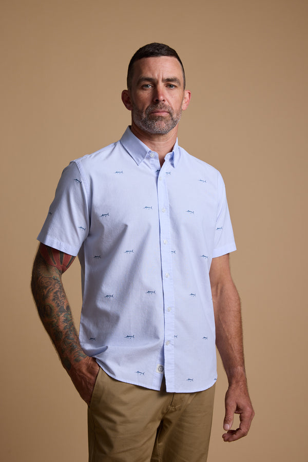 A man with short dark hair, a trimmed beard, and a tattooed right arm wears the Barkers Marlin Stripe Shirt made from responsibly sourced cotton, paired with tan pants, standing against a plain beige background.