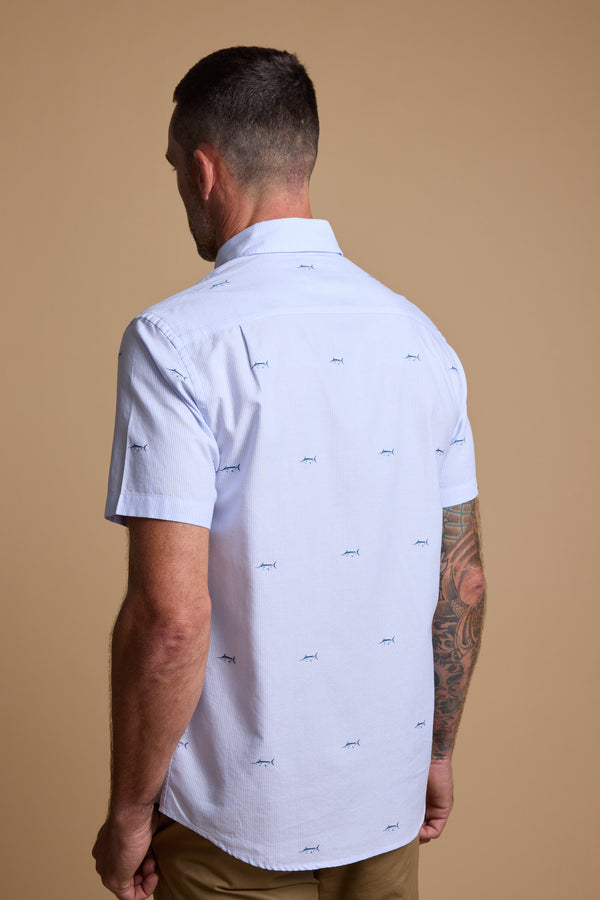 A man with short dark hair and a tattoo sleeve on his left arm, seen from behind, wears the Barkers Marlin Stripe Shirt—light blue, embroidered, made from responsibly sourced cotton—and tan pants against a beige background.