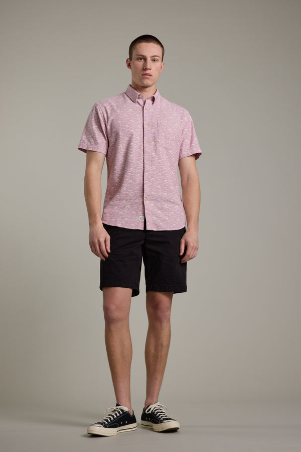 A person stands against a plain background wearing the Barkers Shelby Print Shirt, a light pink short sleeve summer shirt, paired with black shorts and black low-top sneakers with white soles.