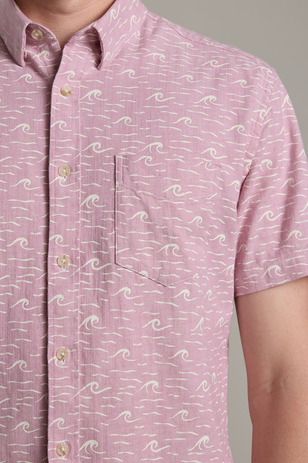 A person wearing the Barkers Shelby Print Shirt—a light pink, short-sleeve summer shirt with a white wave pattern, beige buttons, and a left chest pocket. Made from responsibly sourced cotton. Only the upper torso is visible.