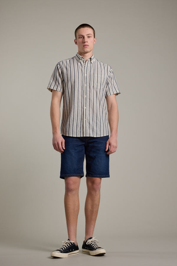 Against a plain light background, a person wears a short-sleeve beige and gray vertically striped shirt, Barkers Rover Denim Short in dark blue, and black low-top sneakers.