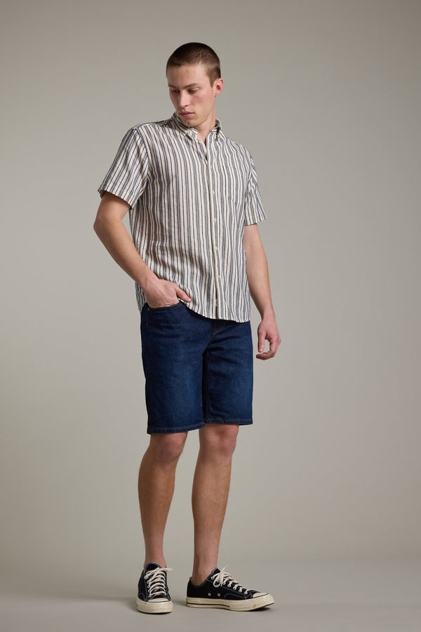 A young person stands against a plain background, wearing Barkers Rover Denim Shorts made from regenerative cotton, a short-sleeve vertical striped shirt, and black sneakers. One hand is in their pocket and their head is slightly turned to the side.