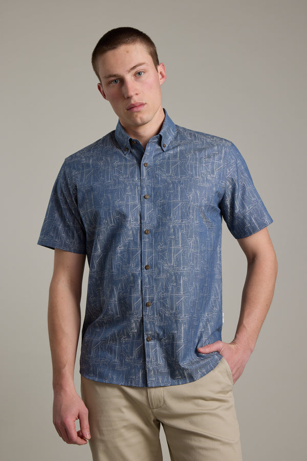 A young man models the Barkers Yacht Jacquard Shirt, a short-sleeved blue summer top, paired with beige pants against a light gray background, one hand in his pocket and wearing a neutral expression.