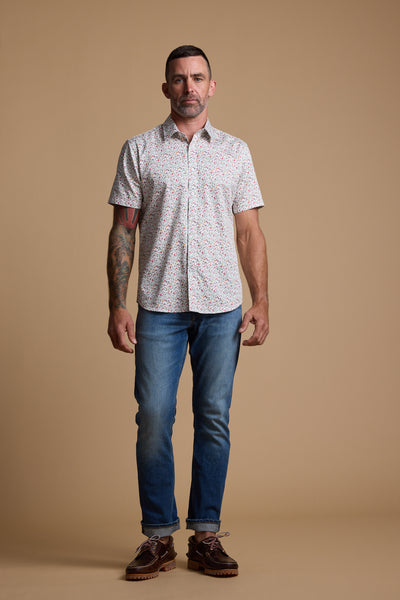 A man with short dark hair and tattoos on his left arm stands against a beige background, wearing the Barkers Wade Print Shirt, blue jeans, and brown shoes.