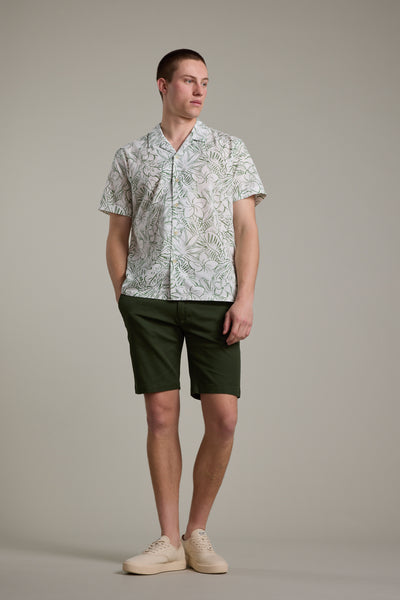A man stands against a plain background wearing a patterned shirt, Braddock Walk Short in dark green by Barkers, and beige sneakers. He looks to the side with one hand in his pocket.
