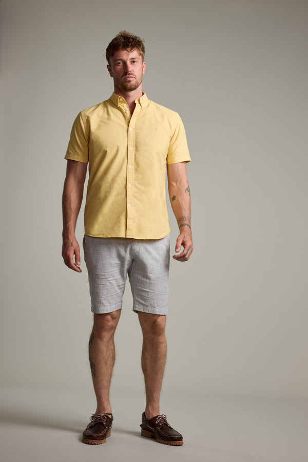 A man faces forward in a Barkers Palma Walkshort, short-sleeve yellow button-up shirt, and brown boat shoes, set against a plain light background.