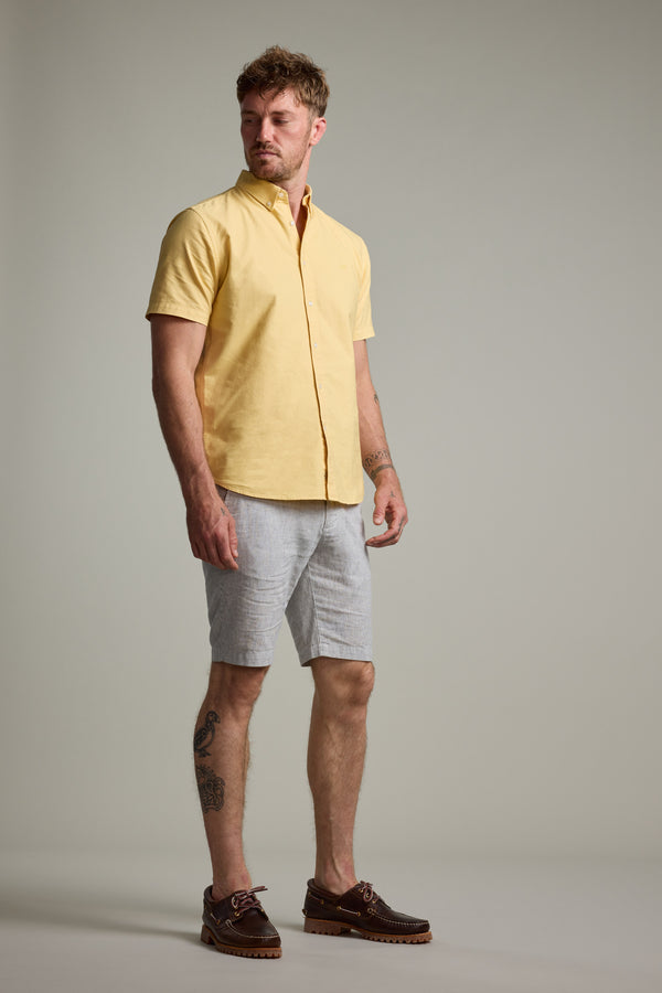 A man stands against a plain background wearing Barkers' Palma Walkshorts in light gray, a short-sleeve yellow button-up shirt, and brown boat shoes. He has short hair, leg tattoos, and looks slightly to the side.