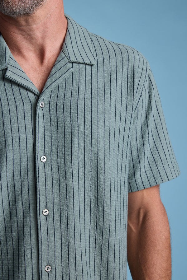 A close-up shows a man in the Barkers Diego Stripe Cuban Shirt—a sage green terry toweling shirt with vertical dark stripes, white buttons, and a boxy fit. Only his lower face, neck, and upper torso are visible against a light blue background.