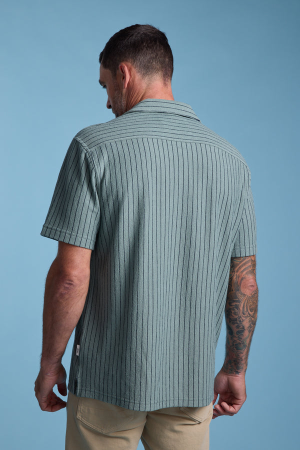 A man with short dark hair and visible arm tattoos models the Barkers Diego Stripe Cuban Shirt, featuring light green fabric with vertical black stripes and a boxy fit, paired with beige pants against a light blue background.