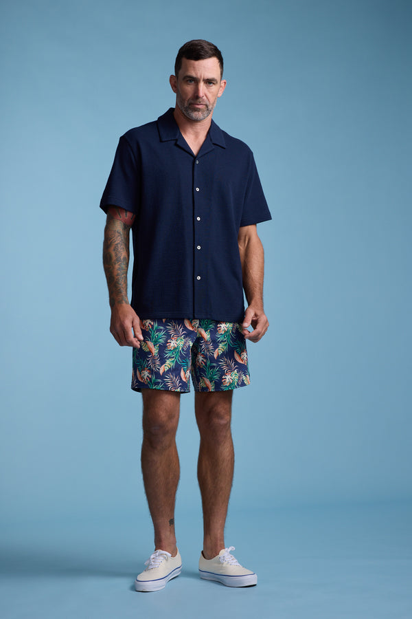 A man with short dark hair and tattoos on his left arm stands against a light blue background wearing the Barkers Diego Cuban Shirt. He pairs it with colorful tropical shorts and white sneakers, sporting a neutral expression.