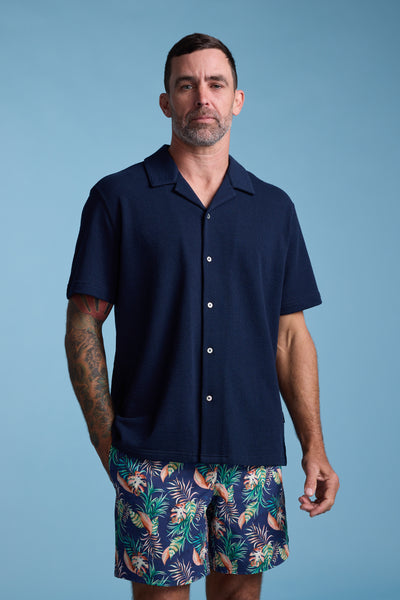 A man with short dark hair, a beard, and tattoos on his left arm stands against a blue background wearing the Barkers Diego Cuban Shirt—a navy short-sleeve shirt—paired with colorful tropical-patterned shorts.