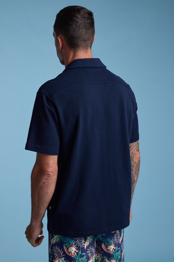 A man with short dark hair, seen from behind, wears a navy Diego Cuban Shirt by Barkers and colorful tropical-print shorts against a solid light blue background.