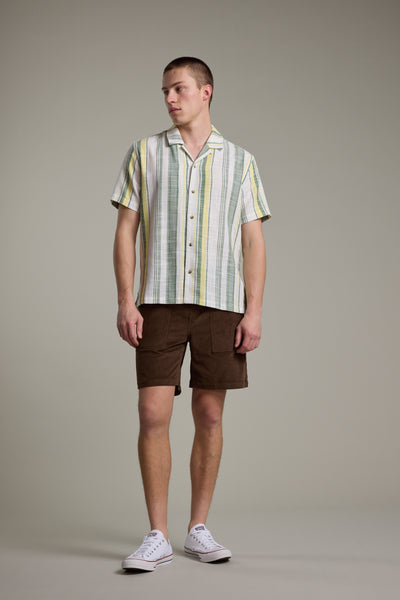 A man stands against a plain background in a short-sleeve, striped button-up shirt with green, yellow, and white tones, Barkers Montauk Cord Weekend Shorts with an elastic waistband, and white sneakers. He looks to the side, relaxed.