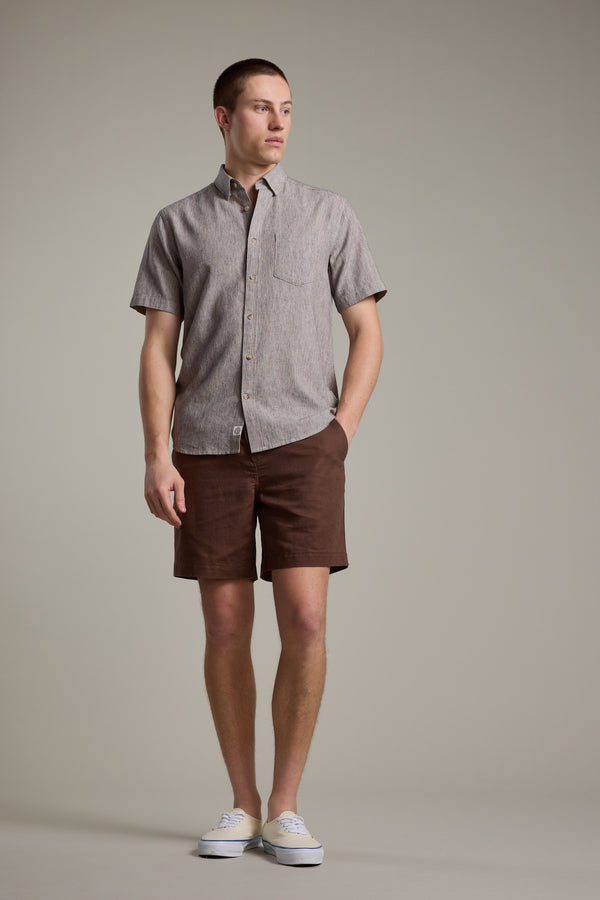 A man stands against a plain background wearing the Barkers Woodley Texture Shirt, styled with brown shorts and white sneakers. The short-sleeve shirt features a cotton-linen blend with thin stripes. He poses with one hand in his pocket, looking to the side.