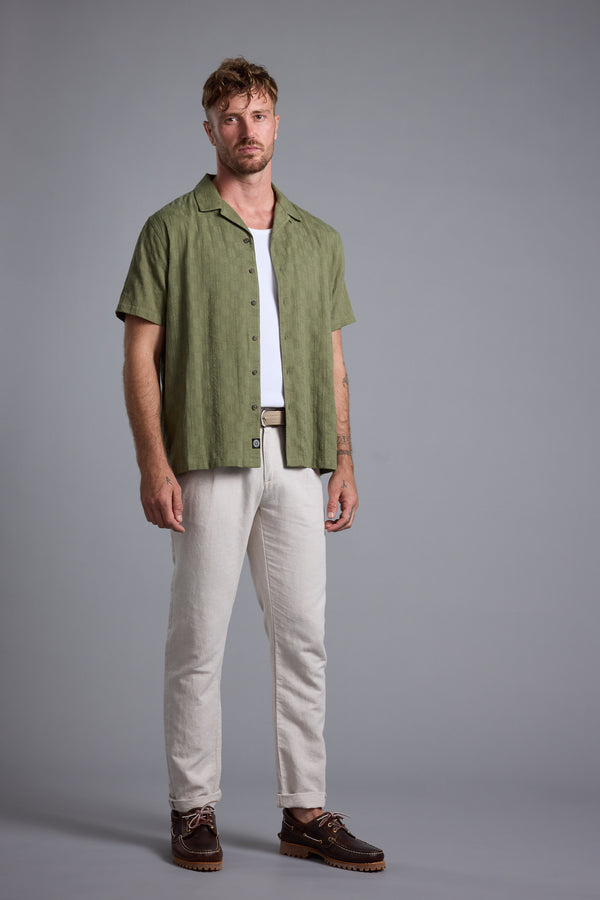 Man in a green Otis Stripe Cuban Shirt by Barkers, white pants & brown shoes, gray background.
