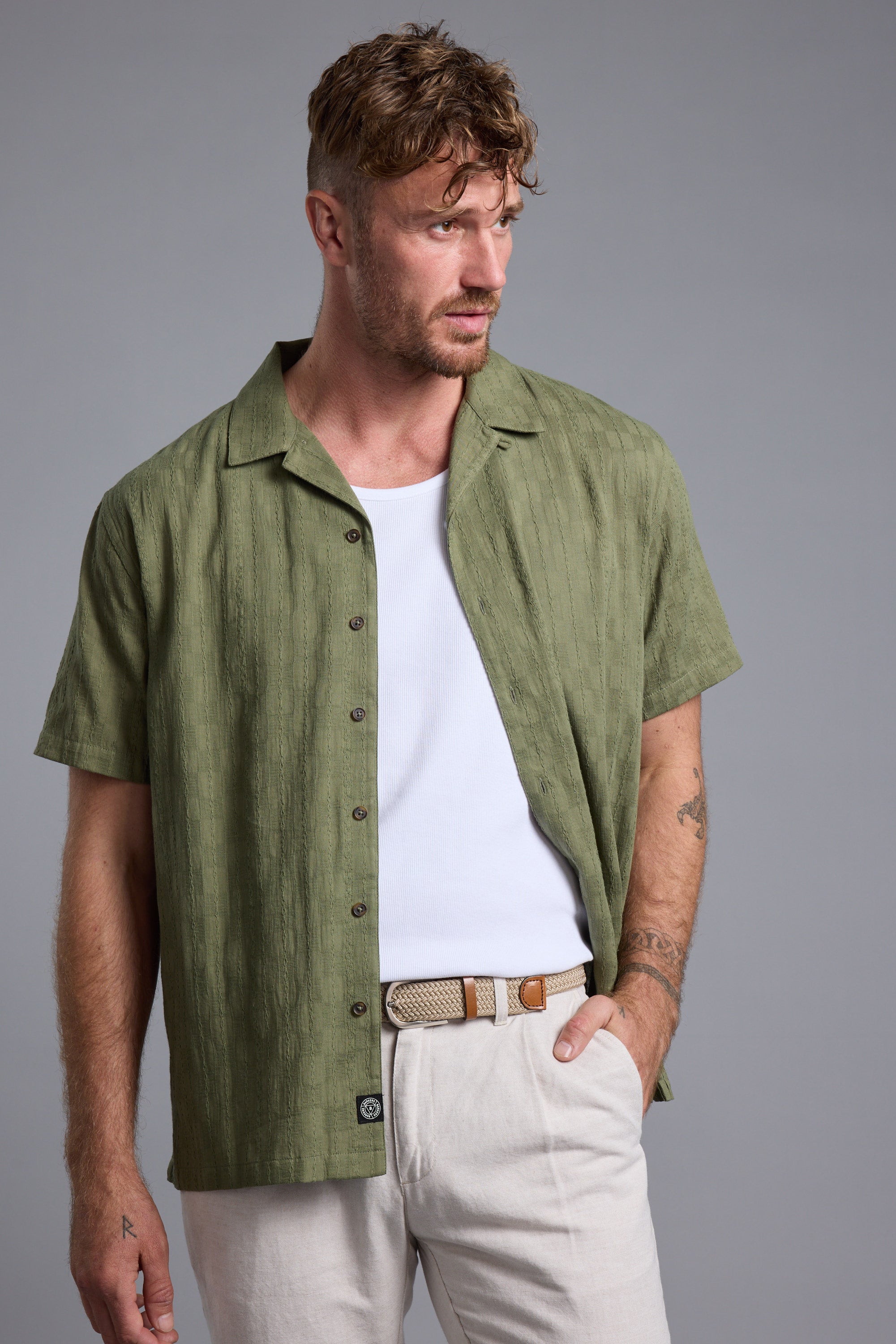 Man in green Otis Stripe Cuban Shirt by Barkers, white tee, beige pants, brown belt, looking left.