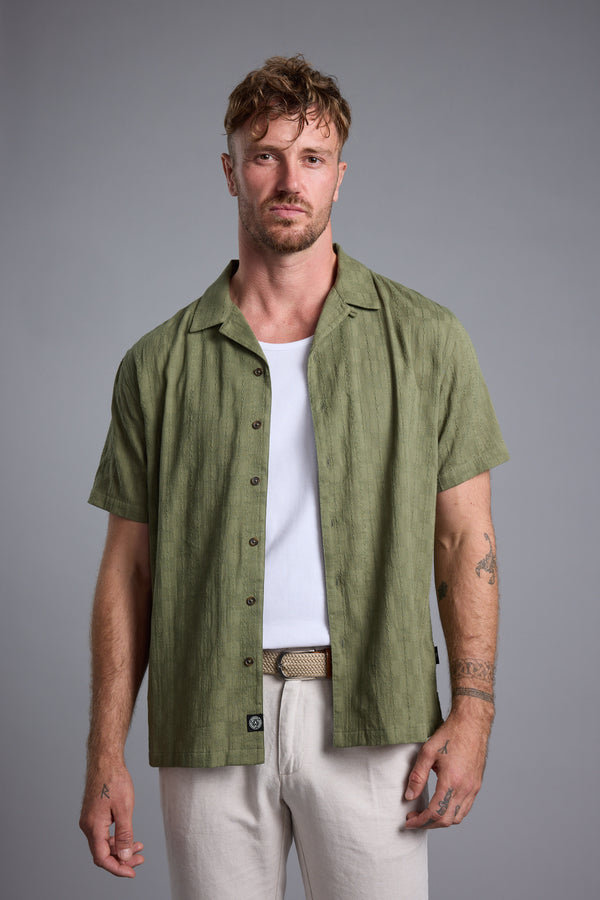 Man in a green Otis Stripe Cuban Shirt by Barkers over a white tank and beige pants, facing forward.