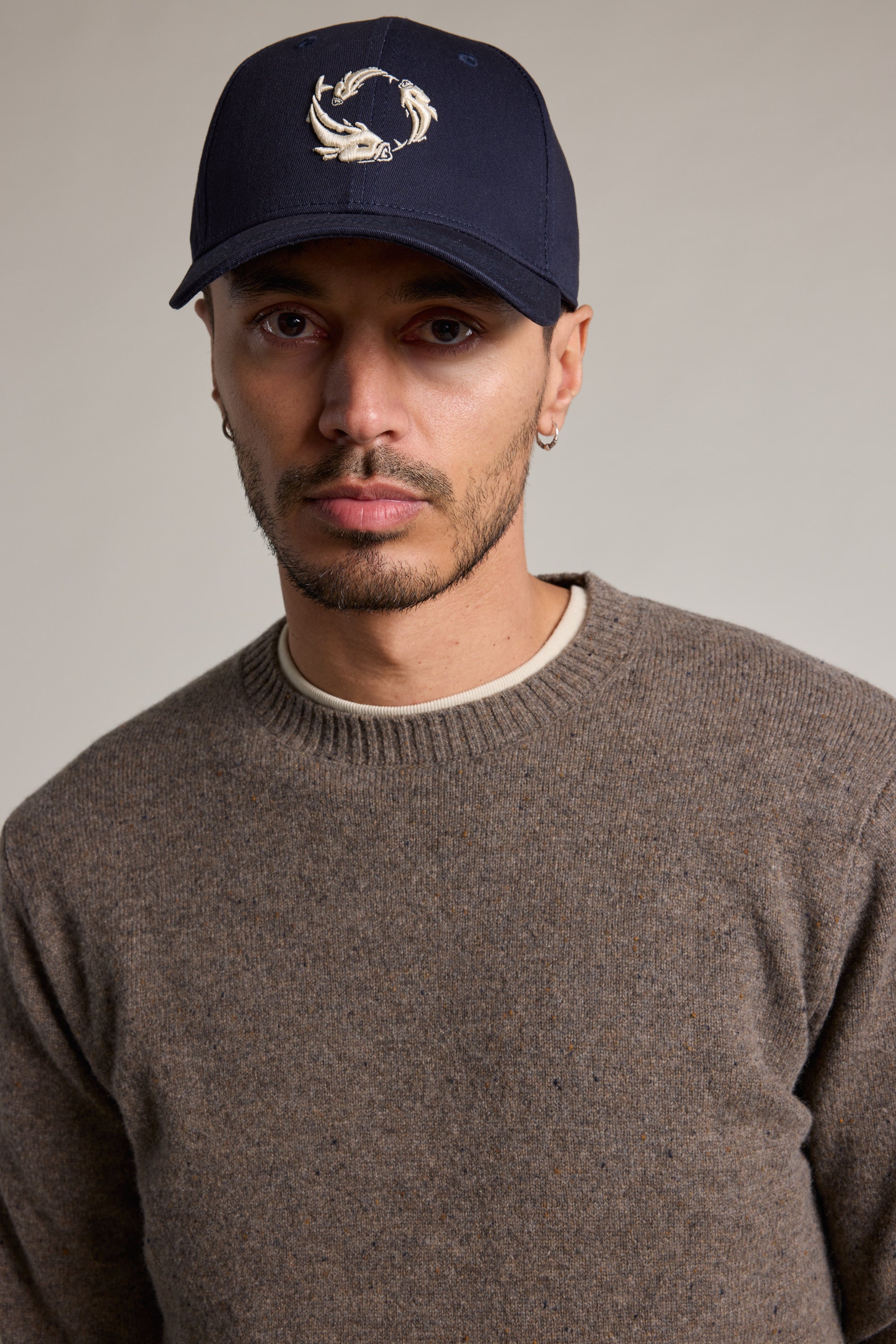 A person with short facial hair wears a navy Legasea Logo Cap by Legasea, with a circular embroidery, brown sweater, and small hoops against a plain background—showcasing New Zealand marine conservation style.