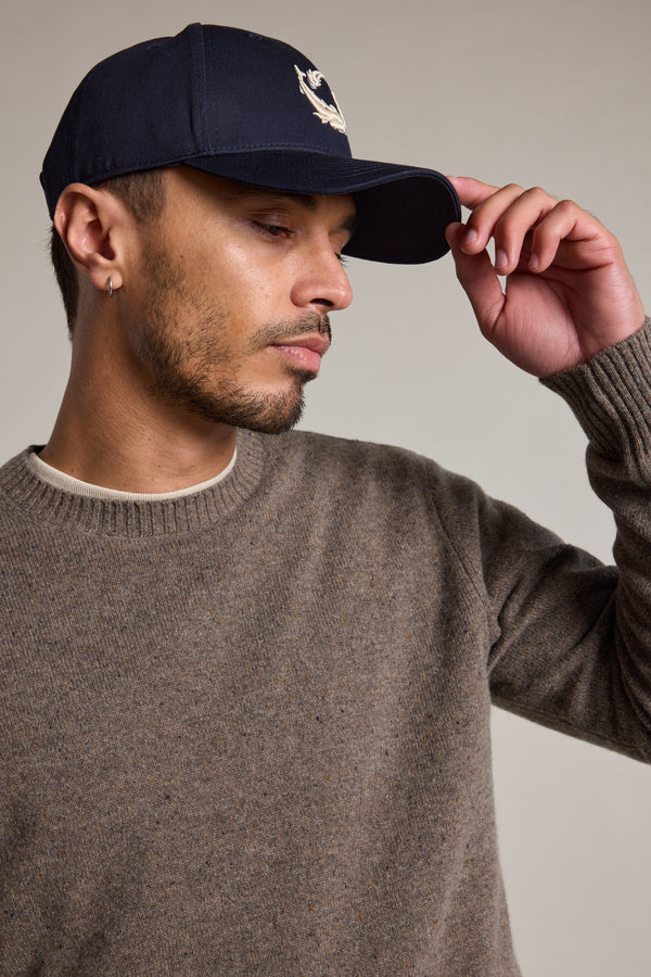 A man in a brown knit sweater wears the Legasea Logo Cap by Legasea, featuring an embroidered fish design, as he tilts the 5-panel cap with one hand and looks down to the side against a plain background.