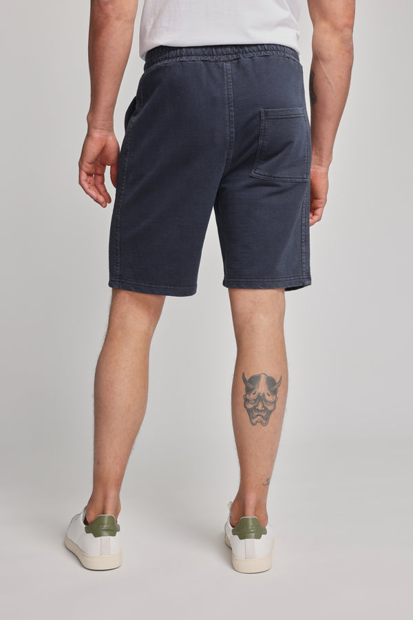A person is photographed from the back, wearing a white t-shirt, Barkers Organic Classic Print Track Short in navy blue with a heritage logo, and white sneakers. A stylized mask tattoo is visible on their right calf.