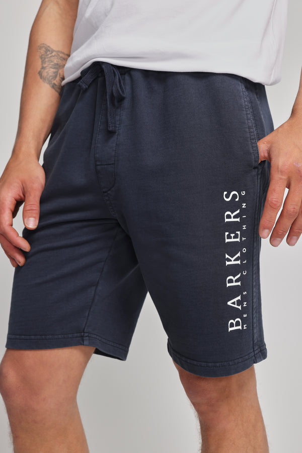 A person wears Barkers Organic Classic Print Track Short in navy, with the heritage logo on the left leg, paired with a white T-shirt. The photo highlights the lower torso and upper legs.