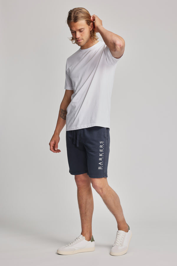 A man with long blond hair in a white t-shirt and navy blue Barkers Organic Classic Print Track Short, featuring the heritage logo, stands against a plain background with one hand touching the back of his head.