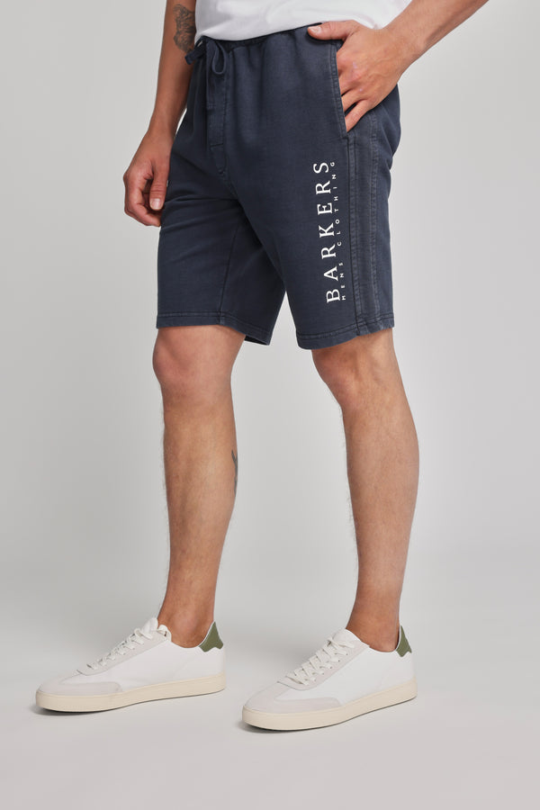 A person wearing navy blue Barkers Organic Classic Print Track Short with the heritage logo on the left leg, paired with a white t-shirt and white sneakers with green accents, stands against a plain light background.