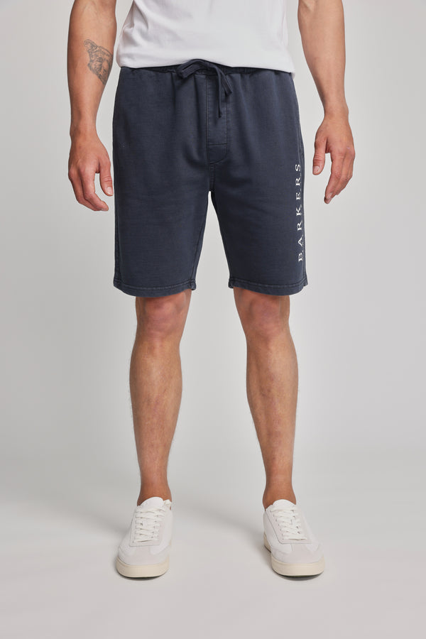 A man, visible only from the waist down, wears Barkers Organic Classic Print Track Shorts in navy with the heritage logo on the left leg, paired with a white T-shirt and white sneakers, standing against a plain light background.