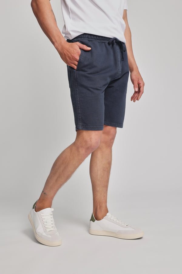 A person wearing a white t-shirt, Barkers Organic Classic Print Track Short in navy, and white sneakers stands with one hand in their pocket against a plain light background.
