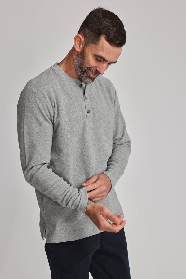 A man with short dark hair and a beard adjusts his sleeve while wearing the Barkers Avenue Waffle Henley in light gray organic cotton and dark pants, set against a plain light background.