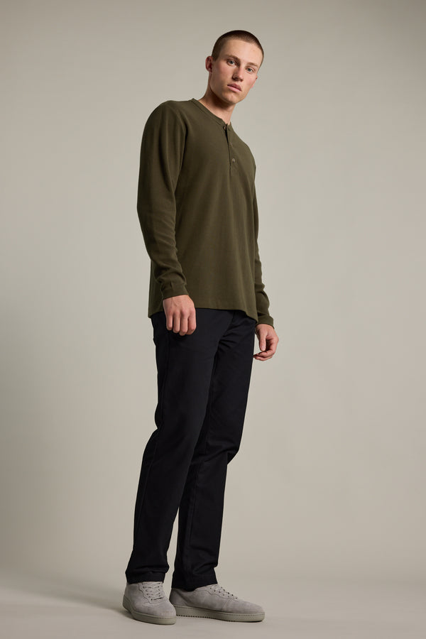 A young man stands against a plain backdrop in the Barkers Avenue Waffle Henley—an olive green organic cotton long-sleeve top—paired with dark pants and light gray sneakers. Ideal as a mid-weight layer for any season, he looks off to the side with a neutral expression.