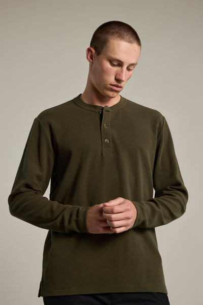 A young man with short hair models the Barkers Avenue Waffle Henley in olive green, made from organic cotton, standing against a light gray background and looking down with his hands loosely clasped in front of him.