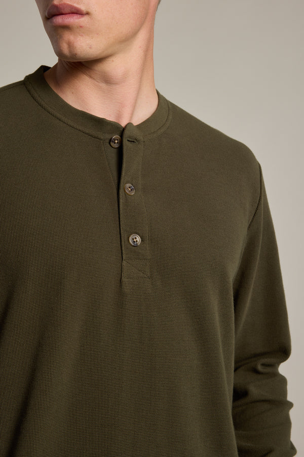 A person wears the Barkers Avenue Waffle Henley in olive green—a mid-weight layer with a three-button collar. The photo captures the upper torso and part of the face against a neutral background.