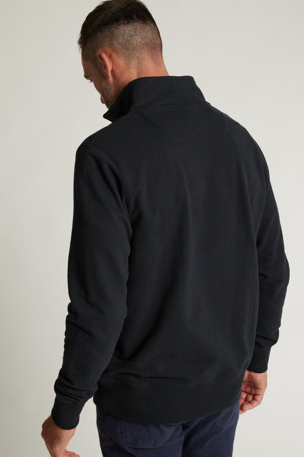 A man with short dark hair stands with his back to the camera, wearing the Barkers Heritage 1/4 Zip sweater by Barkers in black and dark pants against a plain light background.