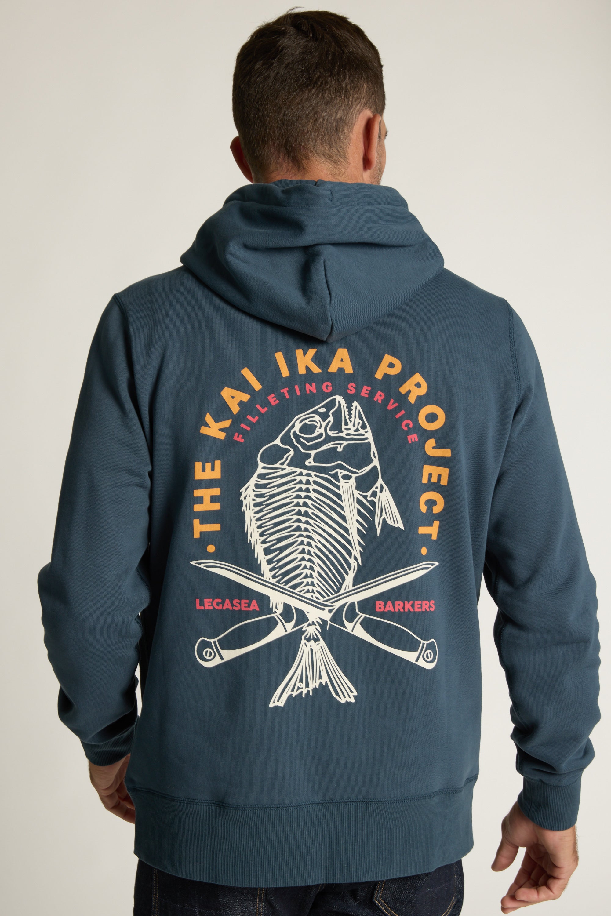 A man wears the Legasea Kai Ika Hoodie by Legasea, an organic cotton blue hoodie featuring a fish skeleton graphic, crossed knives, and THE KAI IKA PROJECT, Filleting Service, LEGASEA, and BARKERS text on the back.