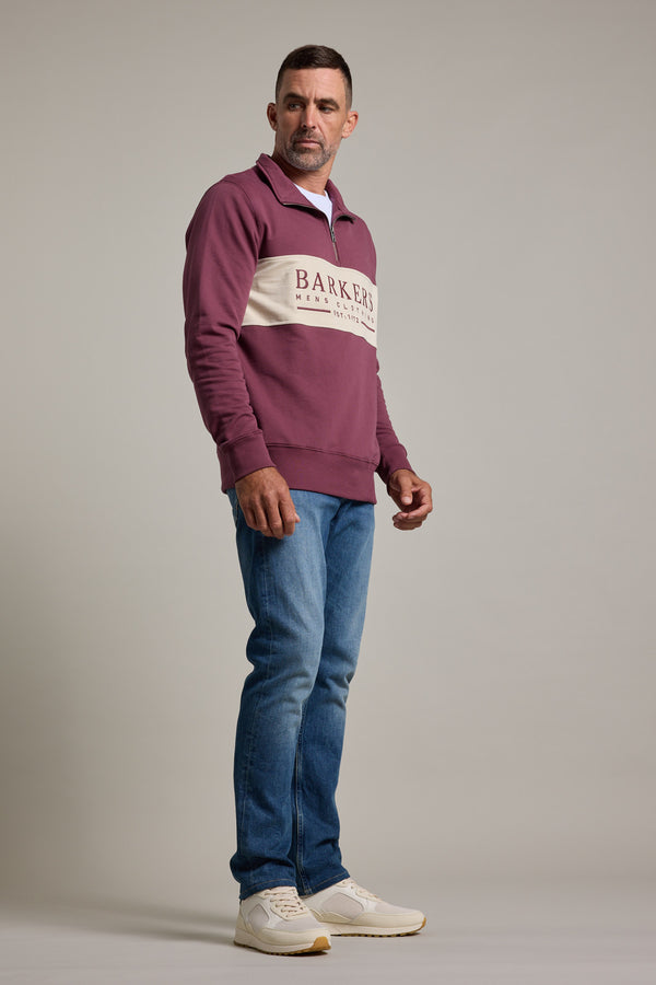 A man stands at an angle wearing the Barkers Heritage 1/4 Zip sweater in mauve by Barkers, featuring a white logo stripe, blue jeans, and white sneakers against a plain light gray background.