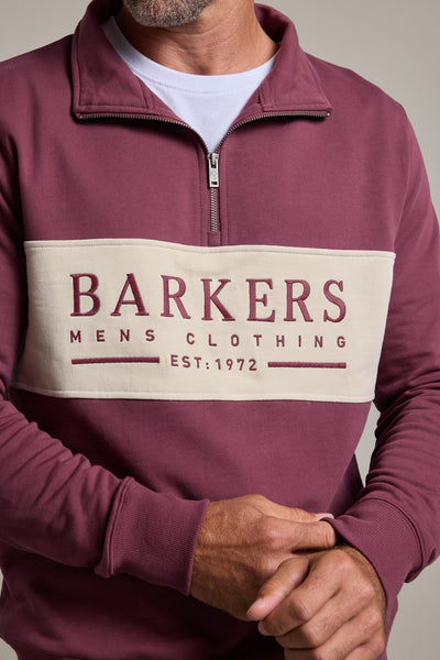 A man wears a maroon and cream Barkers Heritage 1/4 Zip sweater by Barkers, featuring "Barkers Mens Clothing Est: 1972" on the front. Only his torso and hands are visible.