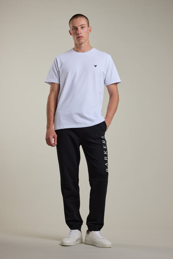 A man with short hair and one hand in his pocket stands against a plain light background, wearing a white t-shirt, Barkers Organic Classic Print Track Pant with BARKERS on the leg, and white sneakers.