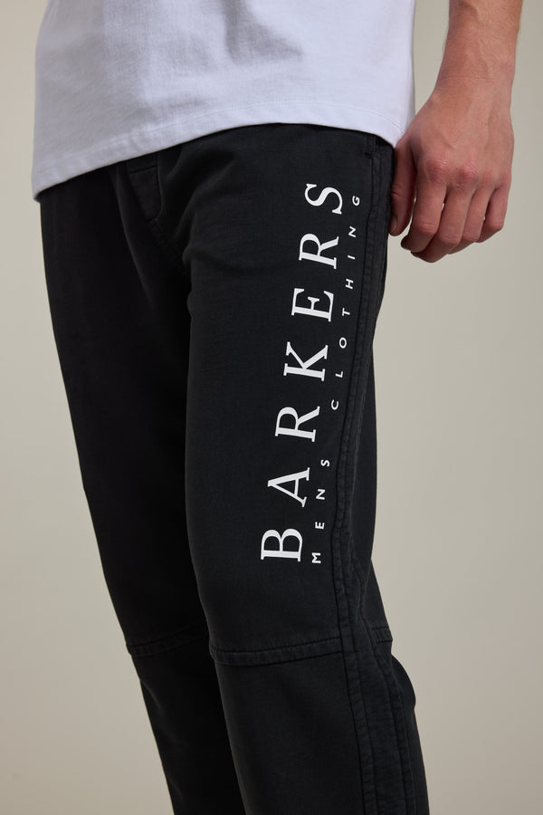 A person wears Barkers Organic Classic Print Track Pant featuring "BARKERS MENS CLOTHING" printed vertically in white on the side, paired with a white shirt. Only the lower torso, right arm, and upper legs are visible.