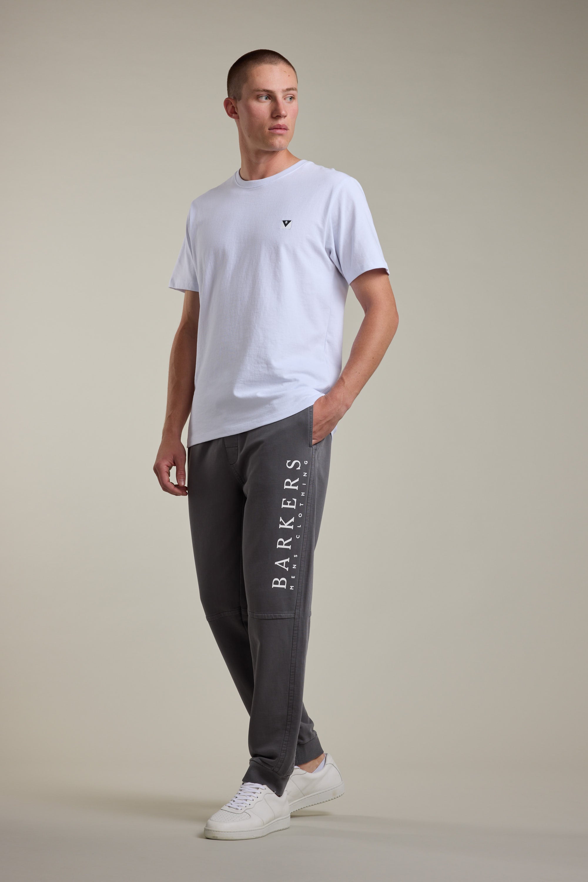 A man stands in a relaxed pose wearing a plain white t-shirt, white sneakers, and Barkers Organic Classic Print Track Pant with BARKERS branding down the leg, against a neutral background.