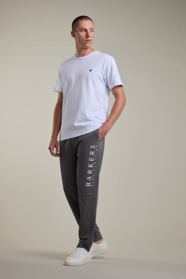 A man stands in a relaxed pose wearing a plain white t-shirt, white sneakers, and Barkers Organic Classic Print Track Pant with BARKERS branding down the leg, against a neutral background.