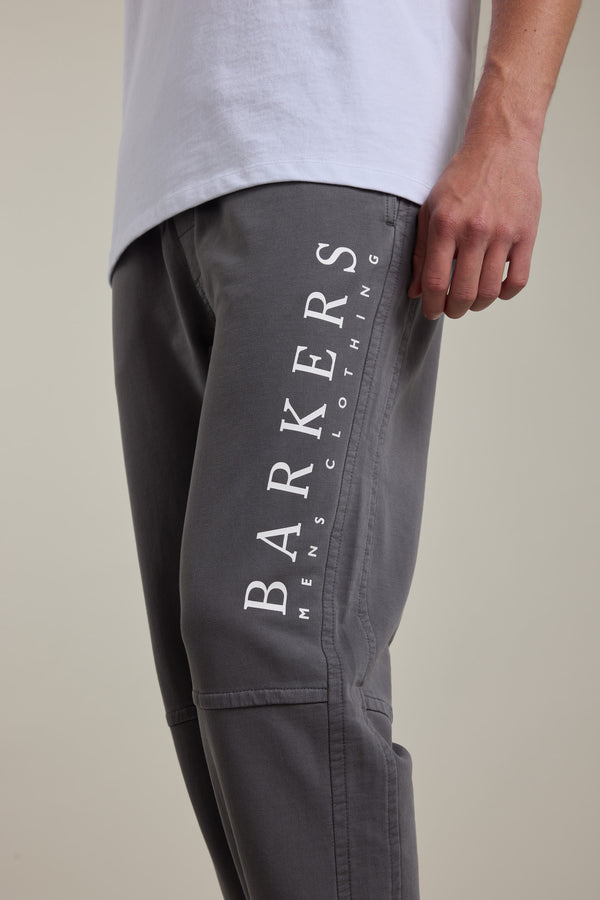 A person is shown from the waist down wearing Barkers Organic Classic Print Track Pant in grey, featuring “BARKERS MENS CLOTHING” printed vertically in white on the thigh, paired with a white shirt.
