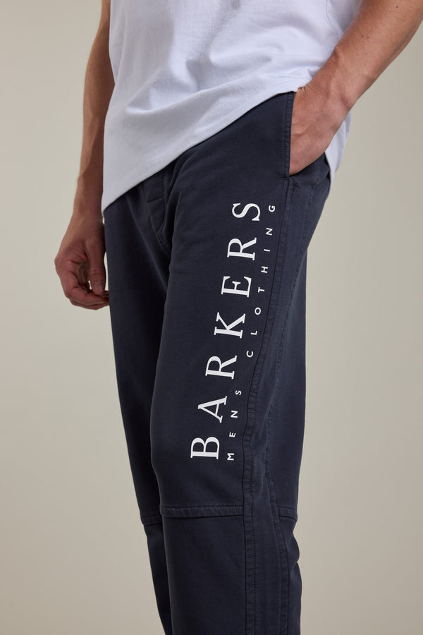 A person wears Barkers Organic Classic Print Track Pant, featuring "BARKERS MENS CLOTHING" vertically on the left thigh, paired with a light t-shirt. Made from organic cotton for comfort, they stand with one hand in their pocket.
