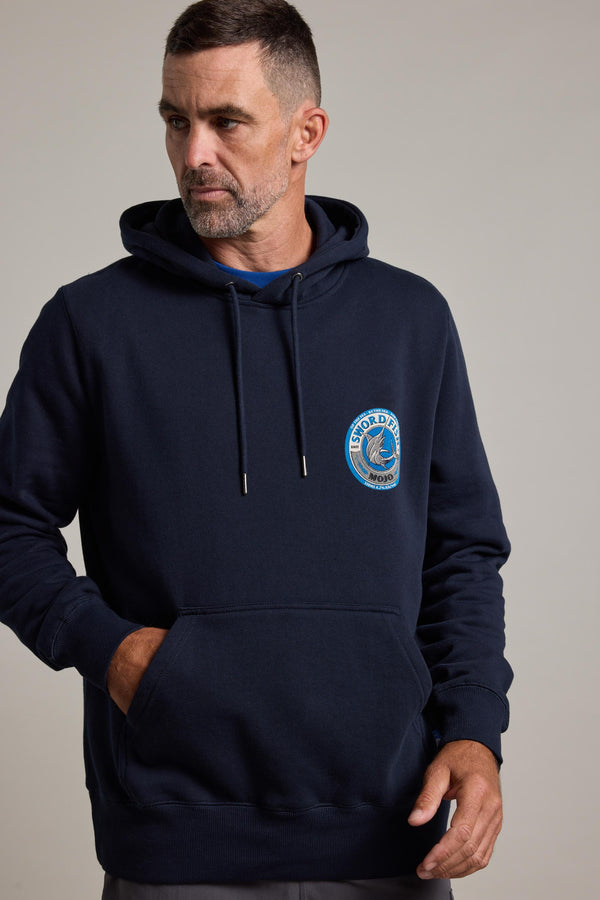 A bearded man with short hair wears the Swordfish Mojo Logo Hoody by Swordfish Mojo in navy blue, made from responsibly sourced cotton. It has a small circular "World" patch on the chest. He looks to the side with one hand in his pocket.