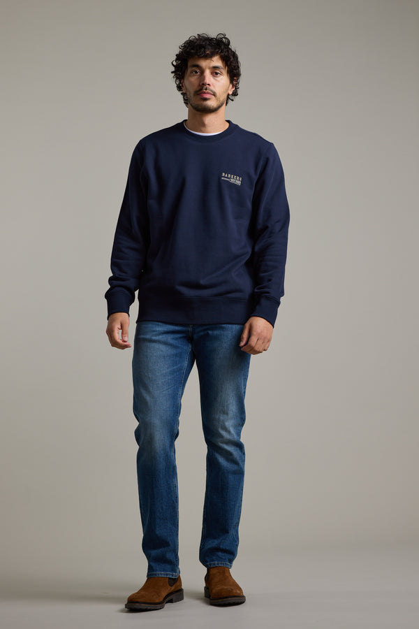 A man with curly hair stands against a plain background, wearing the Barkers Regatta Crew sweatshirt by Barkers, paired with blue jeans and brown suede shoes. He faces forward with a neutral expression.