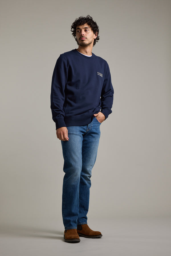 A man with curly hair wears the Barkers Regatta Crew sweatshirt by Barkers, paired with blue jeans and brown boots. He stands against a plain gray background, looking slightly to the side with one hand in his pocket.